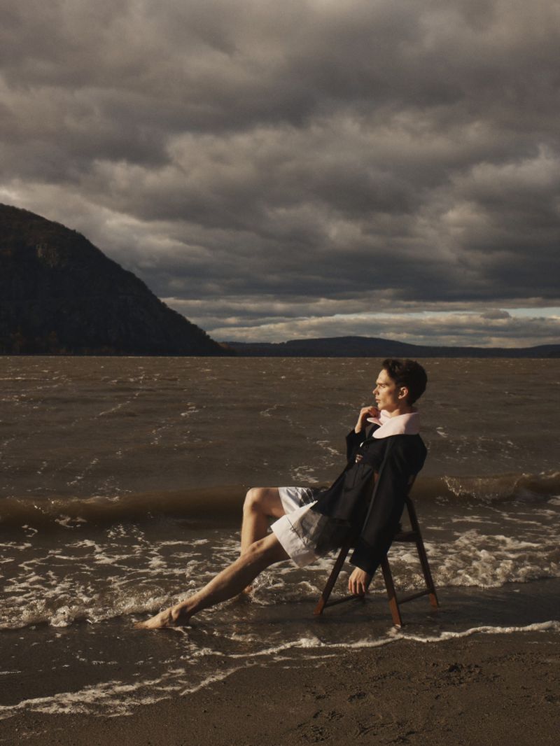 Model seated on a wooden folding chair in shallow water along a sandy shore beneath dark, overcast skies.