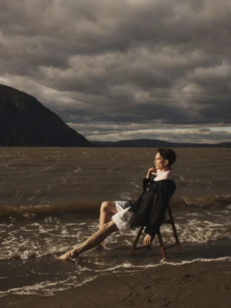 Model seated on a wooden folding chair in shallow water along a sandy shore beneath dark, overcast skies.