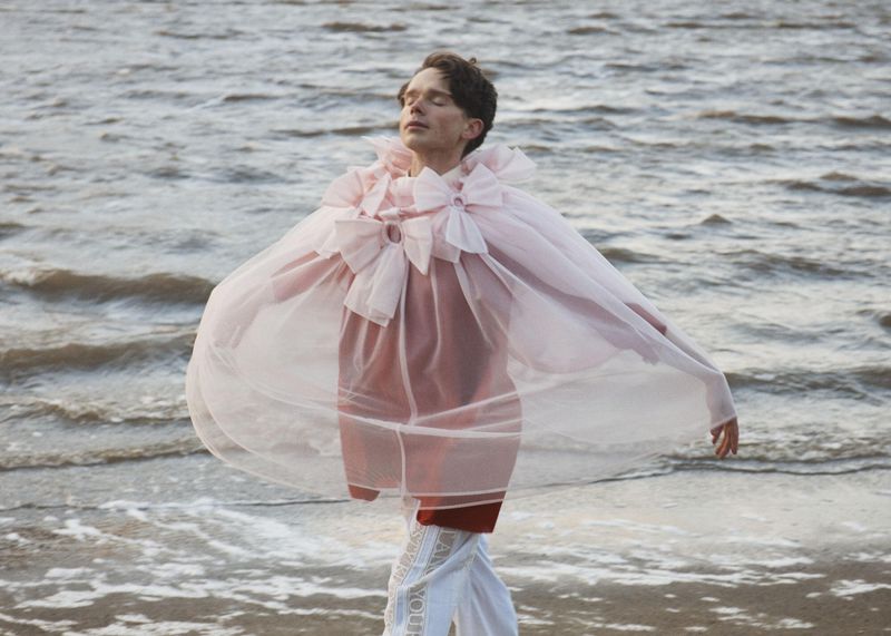 A man wearing a voluminous sheer blush cape with bows standing on a wet sandy shore with ocean waves in the background.