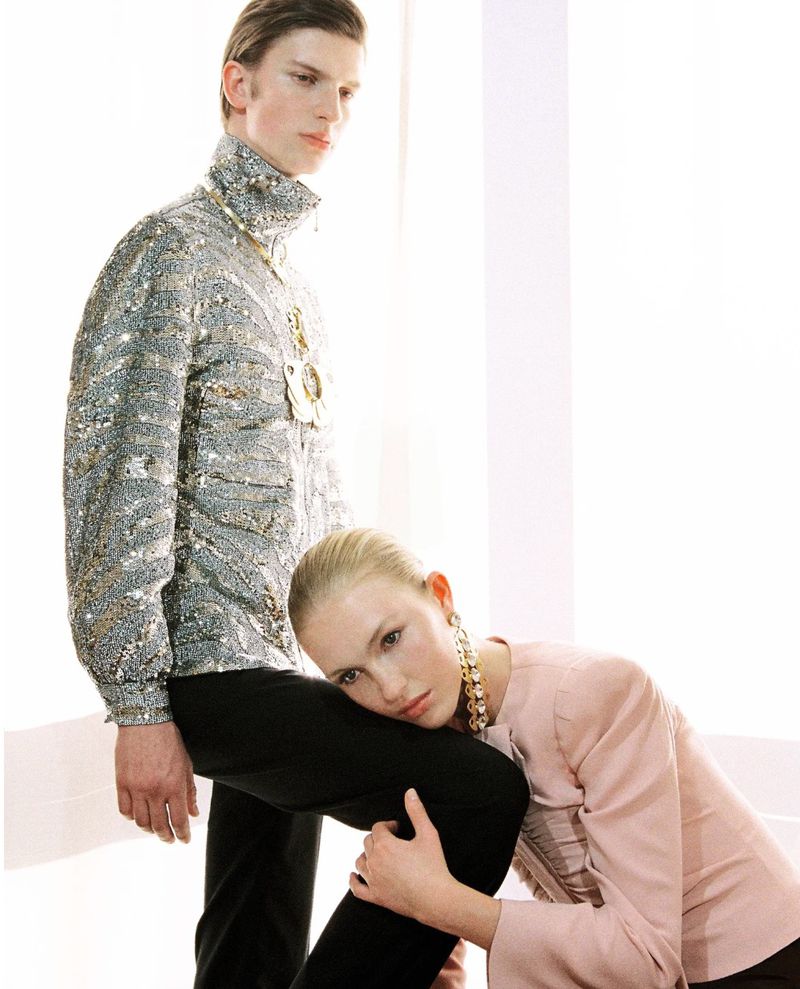 Molly Merland resting her head on a male model's knee wearing a silver sequin jacket in a bright studio.