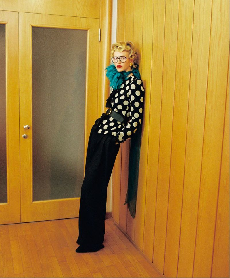 Model Molly Merland in a black and white polka-dot top, wide-leg trousers, and a teal neckpiece leaning against a wood-paneled wall.