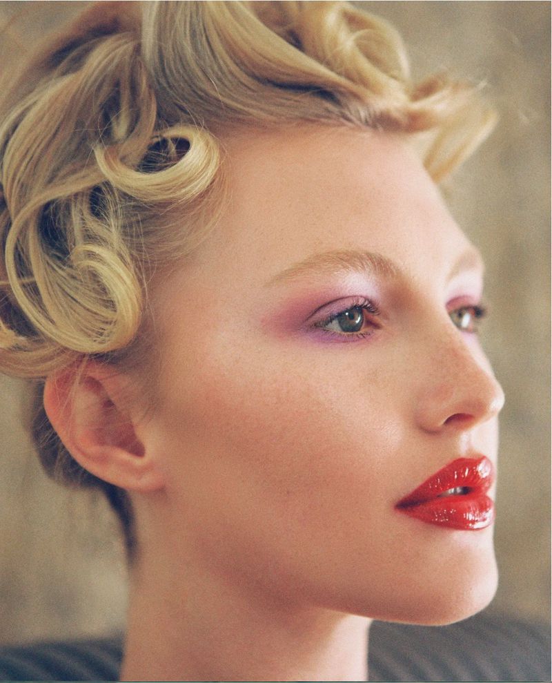 Close-up beauty portrait of a blonde woman with vintage pin curls, purple eyeshadow, and glossy red lipstick.