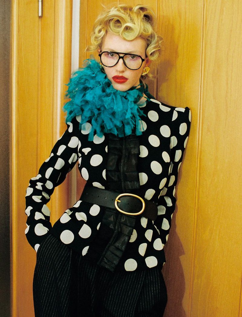 Model Molly Merland wearing black-frame glasses, a teal ruffled collar, and a polka dot jacket against a wooden door.
