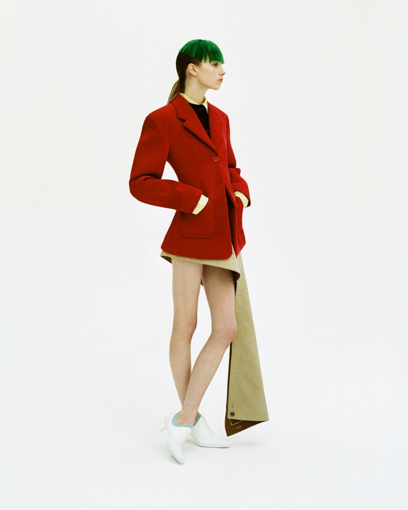 Model with green-dyed fringe wearing a structured red blazer and draped beige skirt against a plain white studio backdrop.