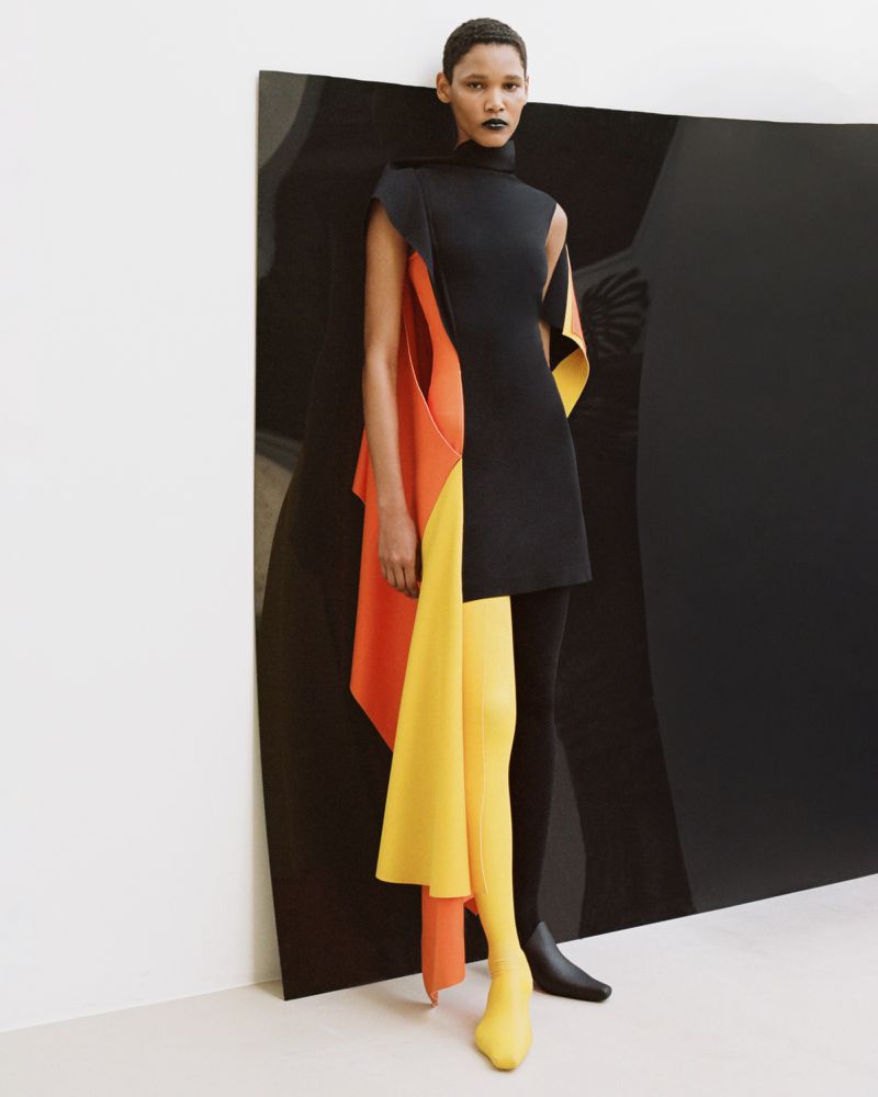 Model wearing a black mini dress with draped yellow and orange panels against a large black curved board in a studio setting.