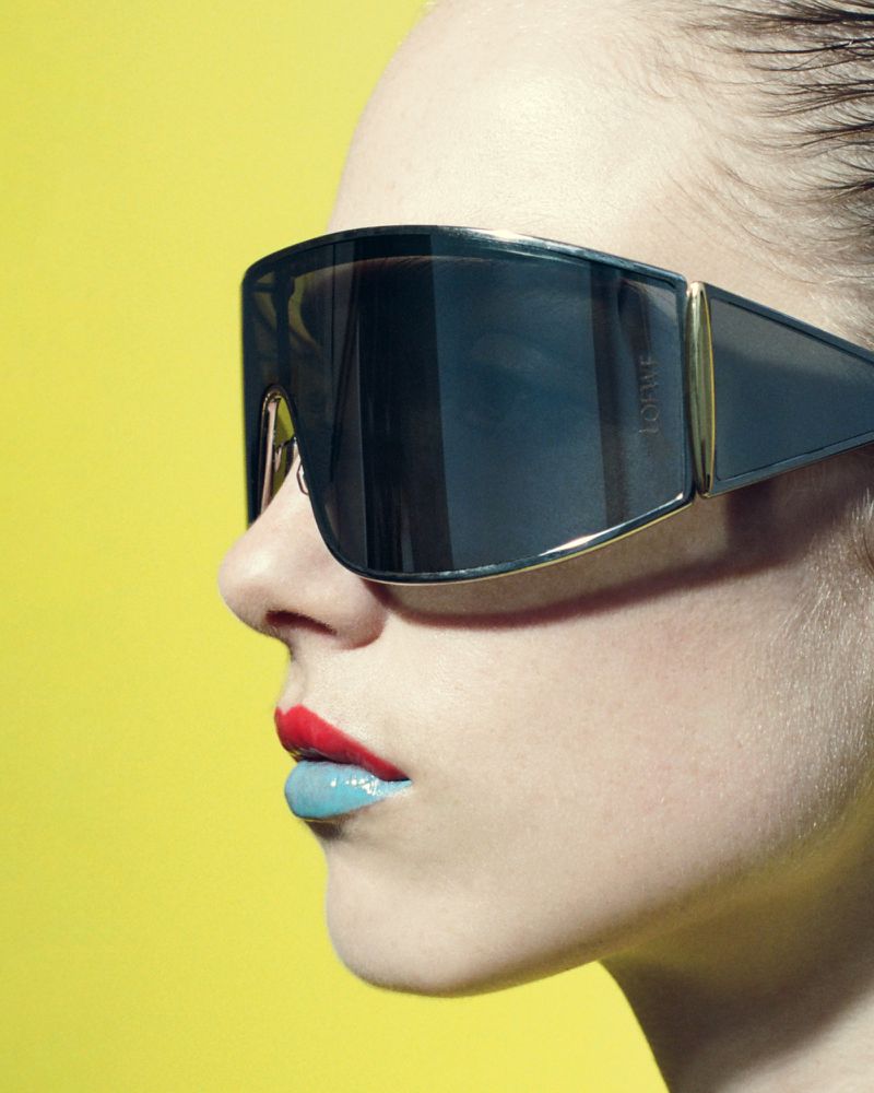Profile close-up of a model wearing black Loewe shield sunglasses with two-tone red and blue lipstick on a yellow background.
