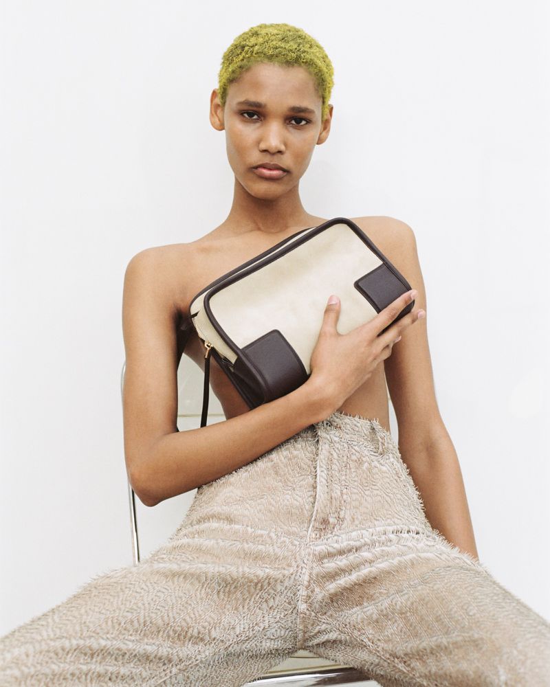 Model with dyed green cropped hair seated holding a cream and brown leather bag wearing textured trousers against a white studio backdrop.