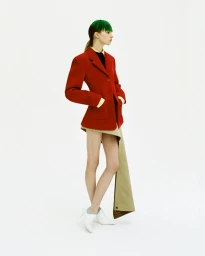 Model with green-dyed fringe wearing a structured red blazer and draped beige skirt against a plain white studio backdrop.