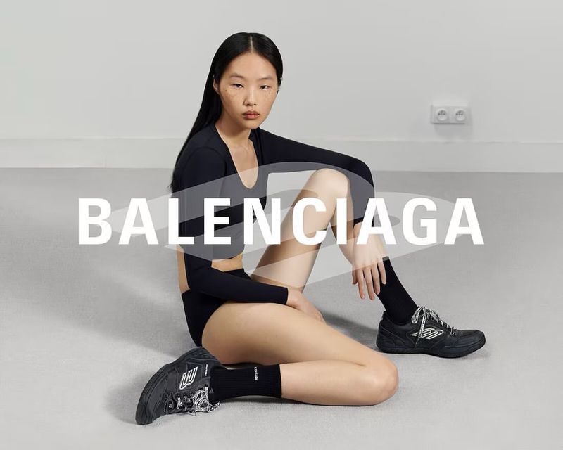 Asian female model sits on the floor wearing a black two-piece outfit and black sneakers, showcasing a Balenciaga advertising campaign.
