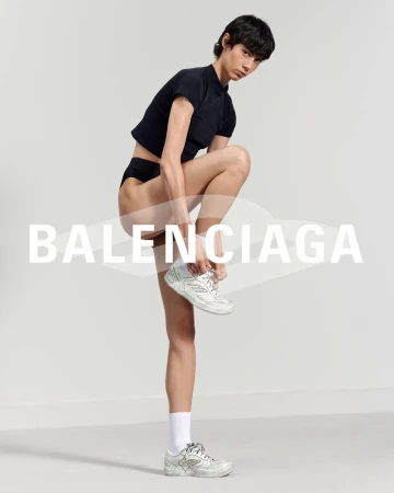 Fashion editorial shot of a slender person with short dark hair wearing a black top, dark underwear, and white Balenciaga sneakers.