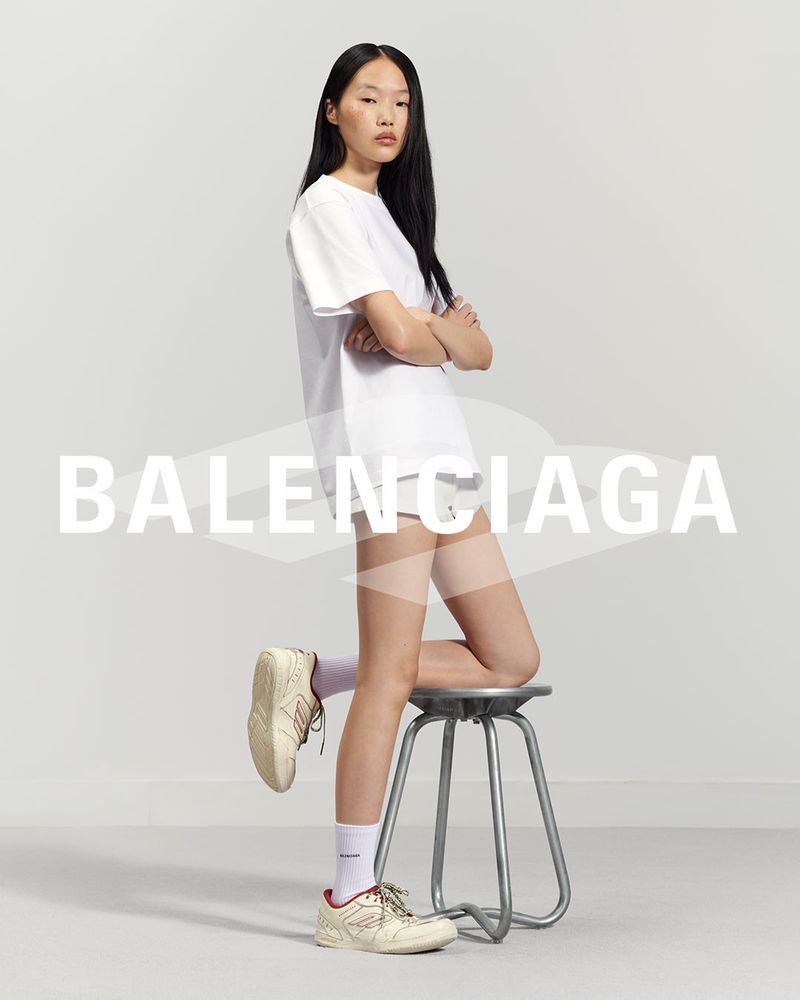 An Asian woman in a white T-shirt and shorts poses against a gray background, with one leg resting on a metal stool.