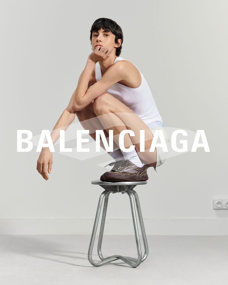 A young adult male poses in a white tank top and brown shoes on a metal stool against a white background with the word BALENCIAGA overlaid.