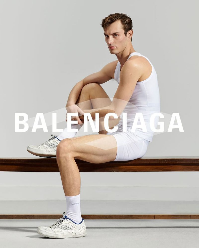 Male model in white tank top and shorts sits on bench with Balenciaga text overlay. Fashion shot features athletic wear and sneakers.