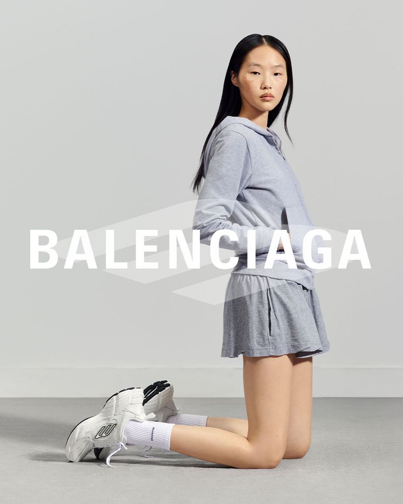 Asian woman posing on her knees in a gray hoodie and shorts, with white sneakers, in a studio setting with the Balenciaga logo.