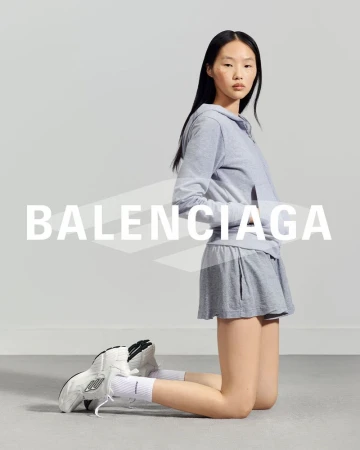 Asian woman posing on her knees in a gray hoodie and shorts, with white sneakers, in a studio setting with the Balenciaga logo.