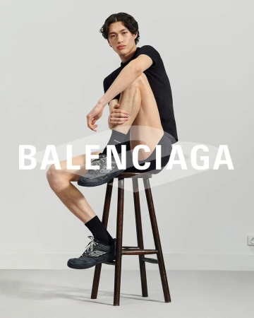 Young adult male seated on a wooden stool in a photo studio, wearing a black t-shirt, athletic shorts, black socks, and Balenciaga sneakers.