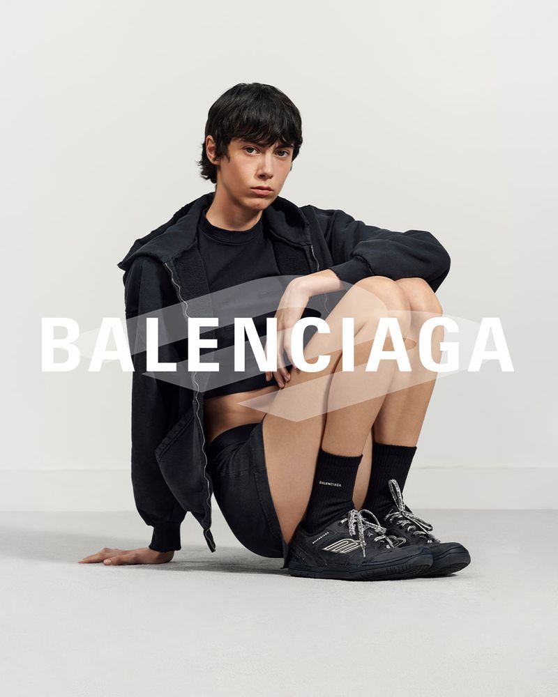 A person sits indoors wearing Balenciaga apparel: a hoodie, t-shirt, shorts, socks, and shoes, with the Balenciaga logo in the foreground.
