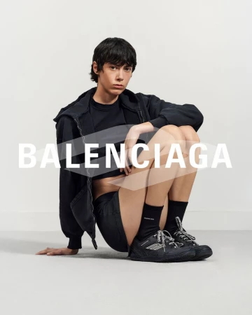 A person sits indoors wearing Balenciaga apparel: a hoodie, t-shirt, shorts, socks, and shoes, with the Balenciaga logo in the foreground.