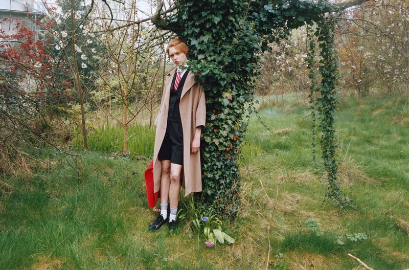 Model Nanne Groenewegen wearing a camel coat and tailored shorts leaning against an ivy-covered tree in an overgrown grassy field.