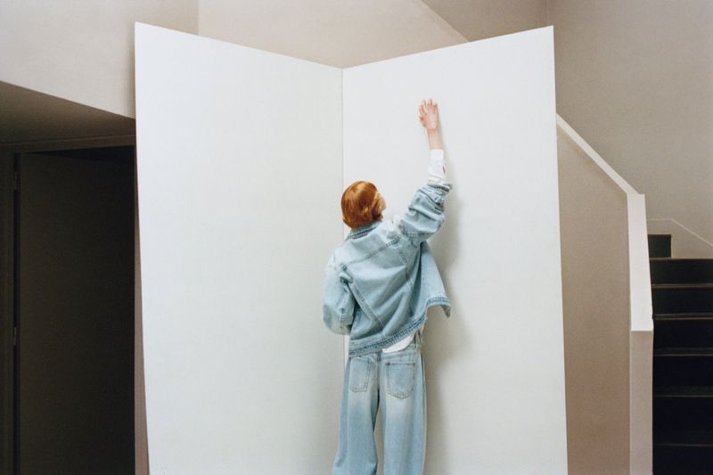 Model Nanne Groenewegen seen from behind in a light wash denim jacket and jeans reaching upward against a white partition backdrop.