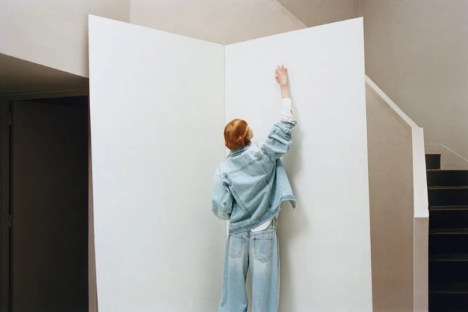 Model Nanne Groenewegen seen from behind in a light wash denim jacket and jeans reaching upward against a white partition backdrop.