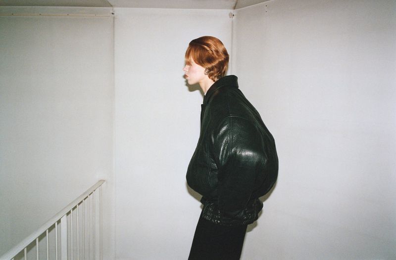 Model in profile wearing an oversized black leather jacket standing near a white railing against plain white walls.