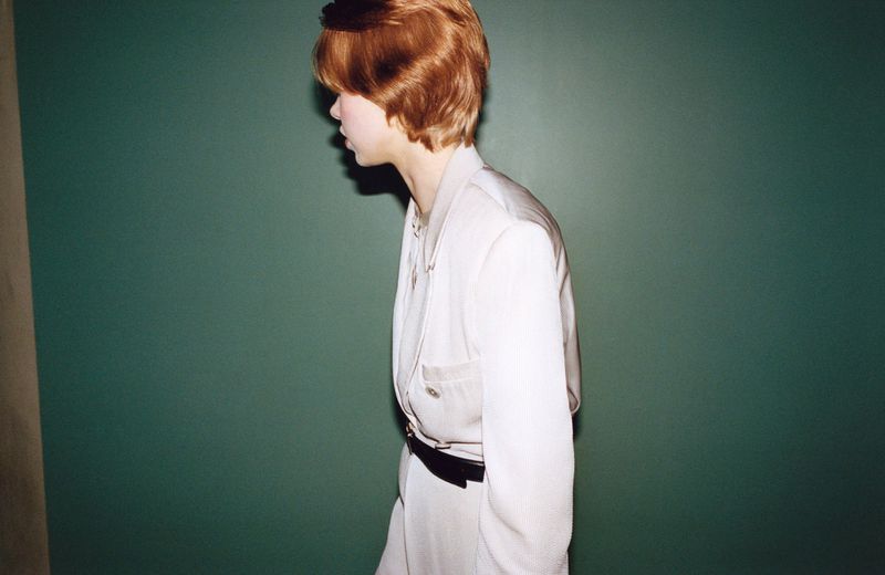 Profile view of model Nanne Groenewegen with short reddish hair wearing an off-white belted suit against a dark green backdrop.