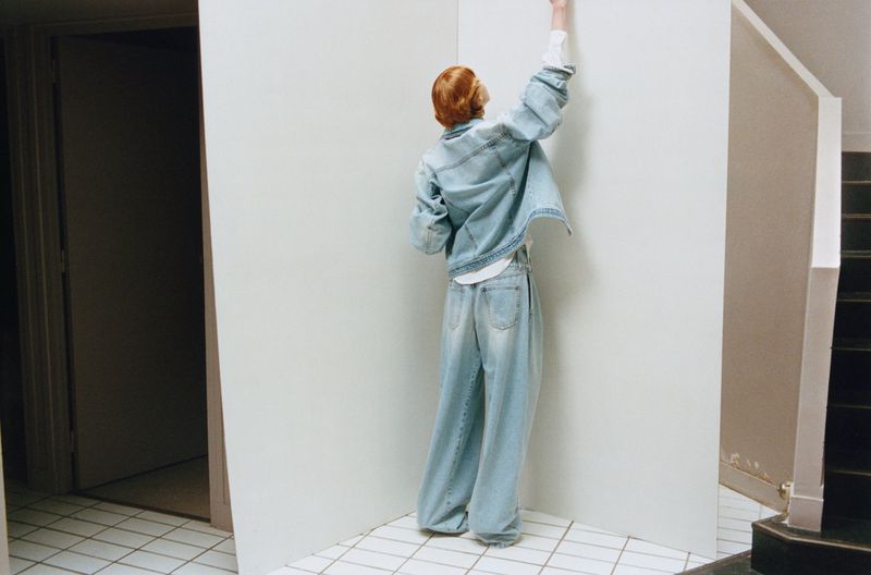 Nanne Groenewegen wearing a light-wash denim jacket and wide-leg jeans, reaching up against a white partition wall.
