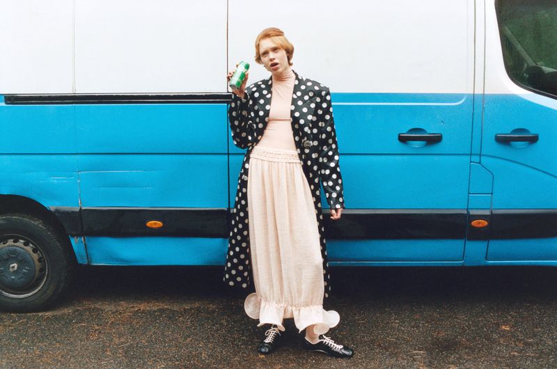 Model Nanne Groenewegen wearing a polka dot coat and ruffled skirt holding a green can in front of a blue van.