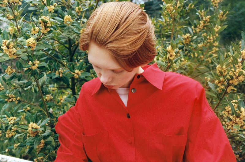 Model Nanne Groenewegen looking downward in a bright red collared shirt with yellow flowering bushes in the background.