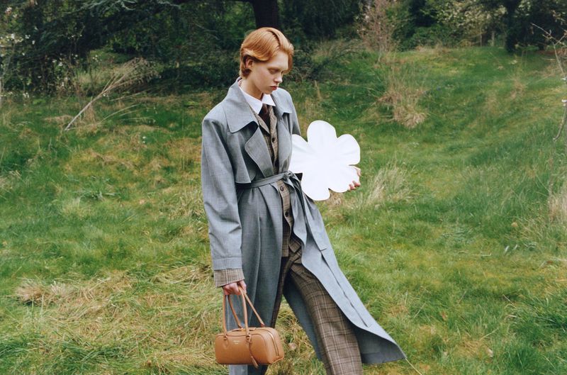 Model Nanne Groenewegen walks across a grassy field in a grey belted trench coat and plaid trousers holding a white flower cutout.