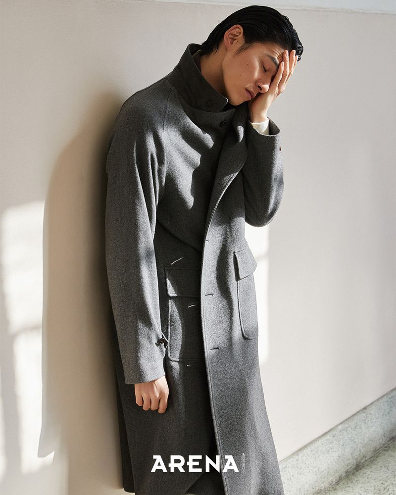 Model Kim Won wearing a long grey wool coat, leaning against a neutral wall with hand to his forehead.