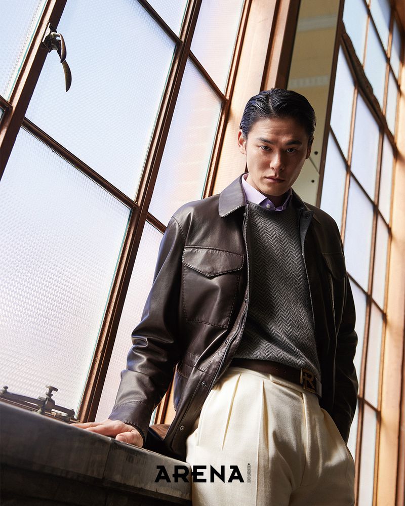Model Kim Won wearing a brown leather jacket, grey sweater, and pleated cream trousers, leaning against a large window.