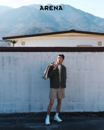 An Asian man stands outdoors wearing a jacket and shorts, a bag over his shoulder, against a wall with mountains in the background.