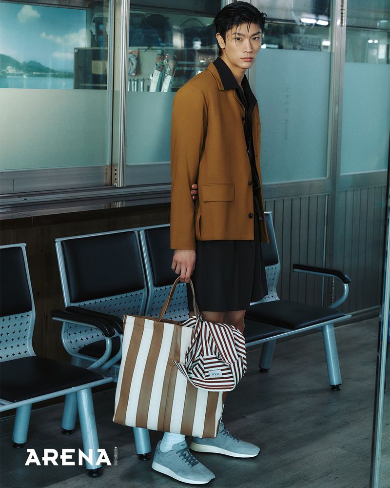 Woohyeong Jang standing by transit waiting chairs in a brown jacket, holding a striped tote bag for Arena Homme+ Korea.