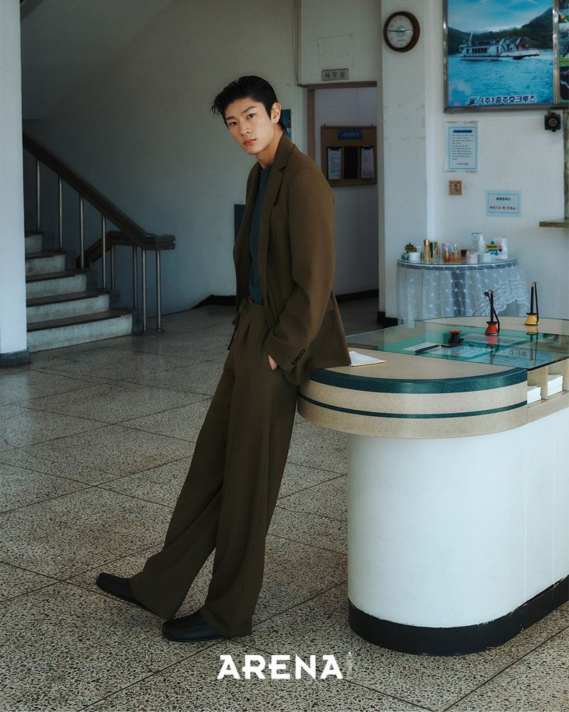 Model Woohyeong Jang posing in an olive brown tailored suit while leaning on a vintage reception desk in a lobby.