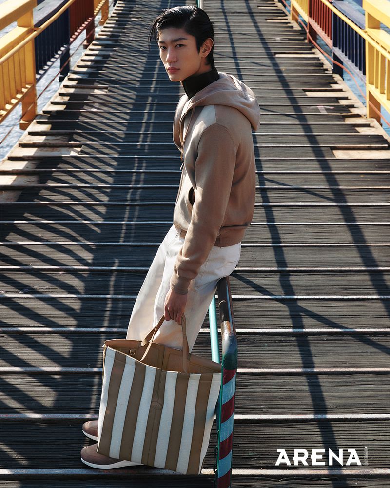 Model Woohyeong Jang wearing a beige hoodie and white pants while holding a striped tote bag on a wooden bridge.