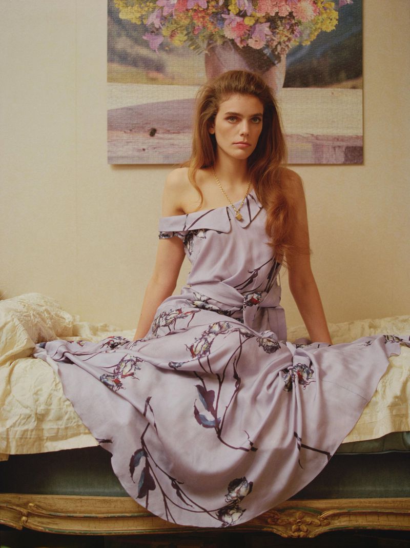 Model Betsy Gaghan seated on a vintage daybed wearing a lilac off-the-shoulder floral dress beneath a floral painting.