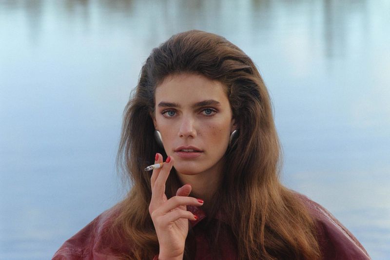 Model with voluminous brown hair holding a lit cigarette, wearing a burgundy jacket and silver earrings by a body of water.