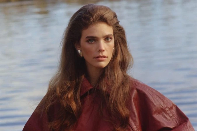 Model Betsy Gaghan wearing a reddish-brown leather jacket and silver earring with rippling water in the background.