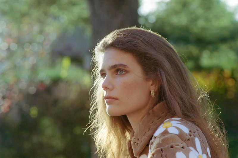 Model Betsy Gaghan in profile wearing a brown daisy-patterned jacket outdoors against soft green foliage.