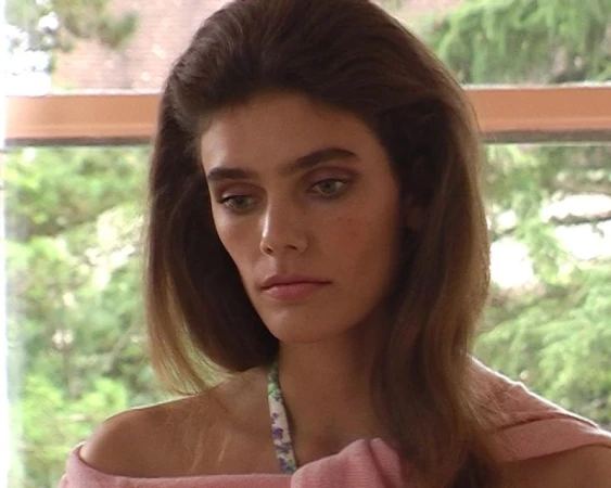 Model Betsy Gaghan with voluminous brunette hair wearing a soft pink off-the-shoulder top in front of a window overlooking greenery.