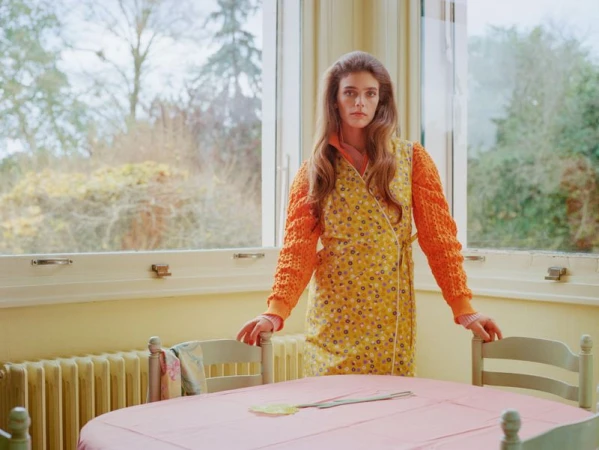 Model Betsy Gaghan wearing an orange knit sweater and yellow floral wrap dress standing behind a dining table by a large window.