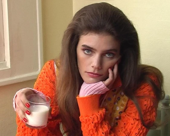 Model with voluminous brunette hair wearing an orange knit sweater and holding a glass of milk.