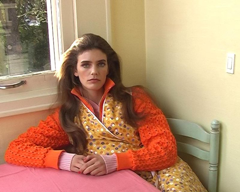 Model Betsy Gaghan seated at a pink table wearing an orange knit sweater and patterned dress beside a window in a corner room.