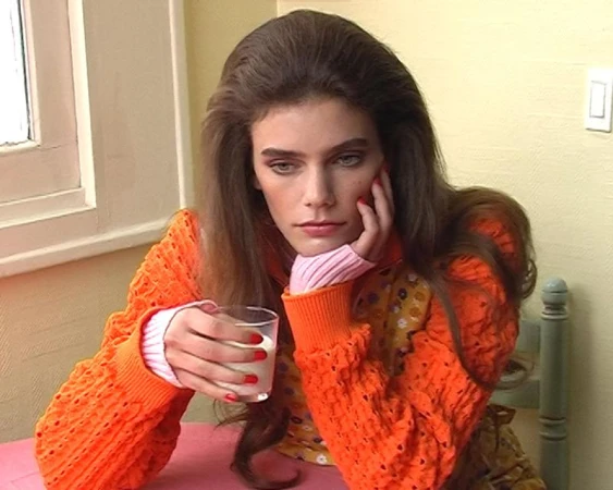 Betsy Gaghan in an orange textured knit sweater resting her chin on her hand and holding a glass of milk at a table.