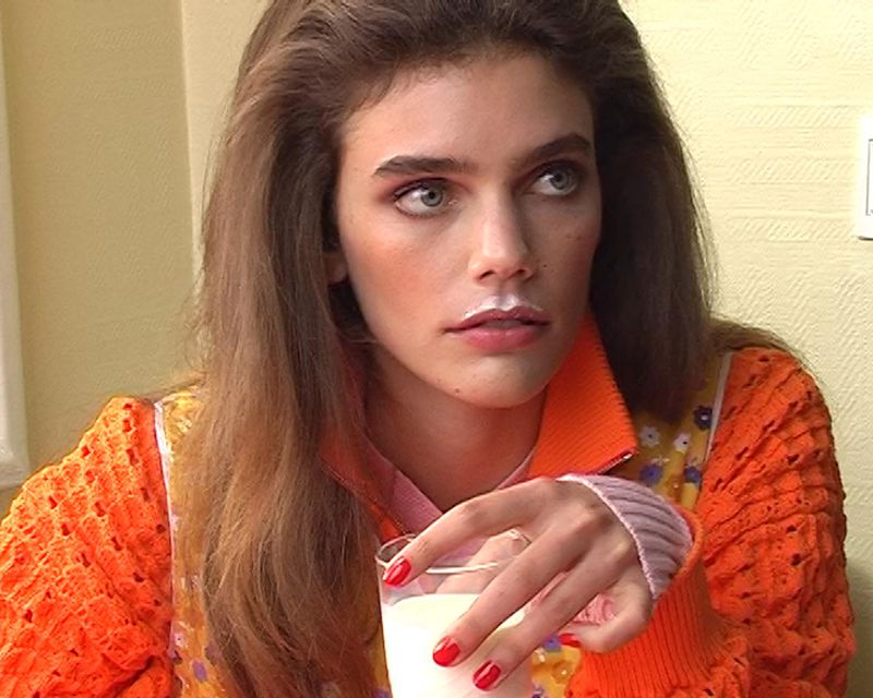 Model Betsy Gaghan with a milk mustache holding a glass of milk while wearing a vibrant orange knit top.