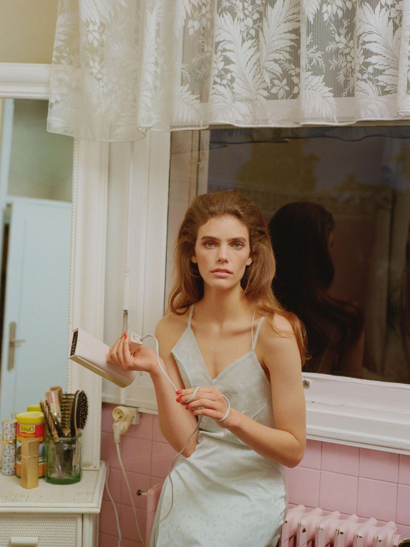 Model Betsy Gaghan in a pale slip dress holding a styling device in a vintage bathroom setting with pink tiles and lace curtains.