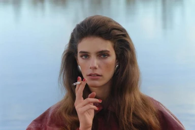 Model with voluminous brown hair holding a lit cigarette, wearing a burgundy jacket and silver earrings by a body of water.