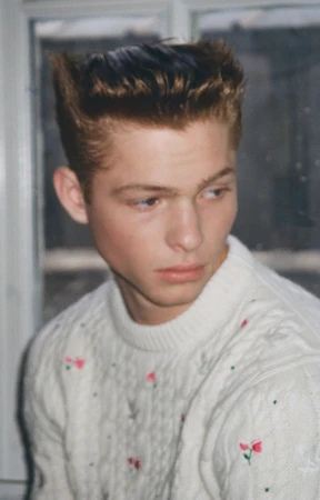 Jasper Hodges wears an off-white embroidered cable-knit sweater posing indoors in front of a window pane.